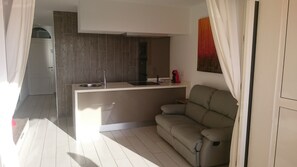 Una cocina moderna con un lavamanos integrado y una pequeña isla, un sofá beige y un comedor con una silla roja.