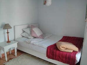 A single-bed room with a white headboard, a red blanket, and a small bedside table with a lamp.