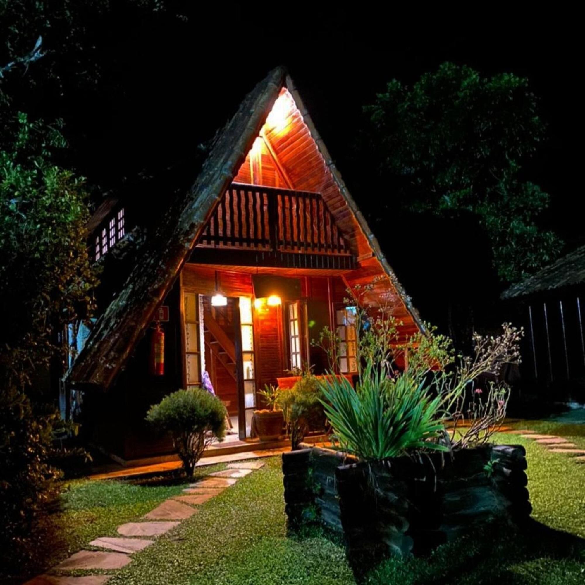 Uma casa de madeira triangular com varanda, iluminada por uma luz quente.