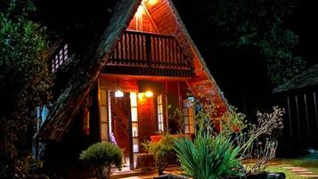 Uma casa de madeira triangular com varanda, iluminada por uma luz quente.