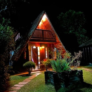 A triangular wooden house with a porch, illuminated by warm lighting. - pousada recanto das aves (Iguape)