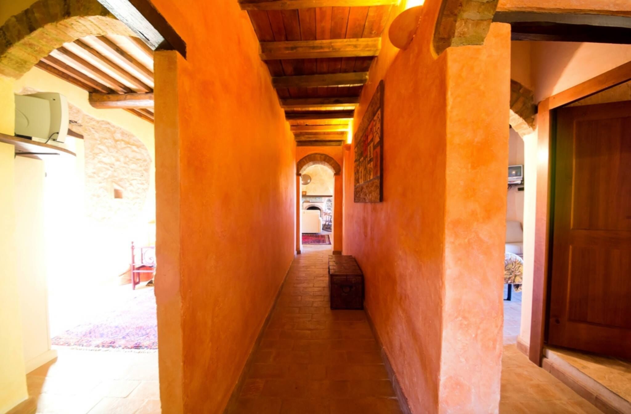 Un estrecho pasillo con vigas de madera a la vista y paredes de piedra, que conduce a una habitación con suelo de baldosas y una puerta.