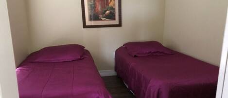 Una habitación con dos camas individuales, cada una con una colcha morada, un cuadro en la pared y una puerta visible a la derecha.
