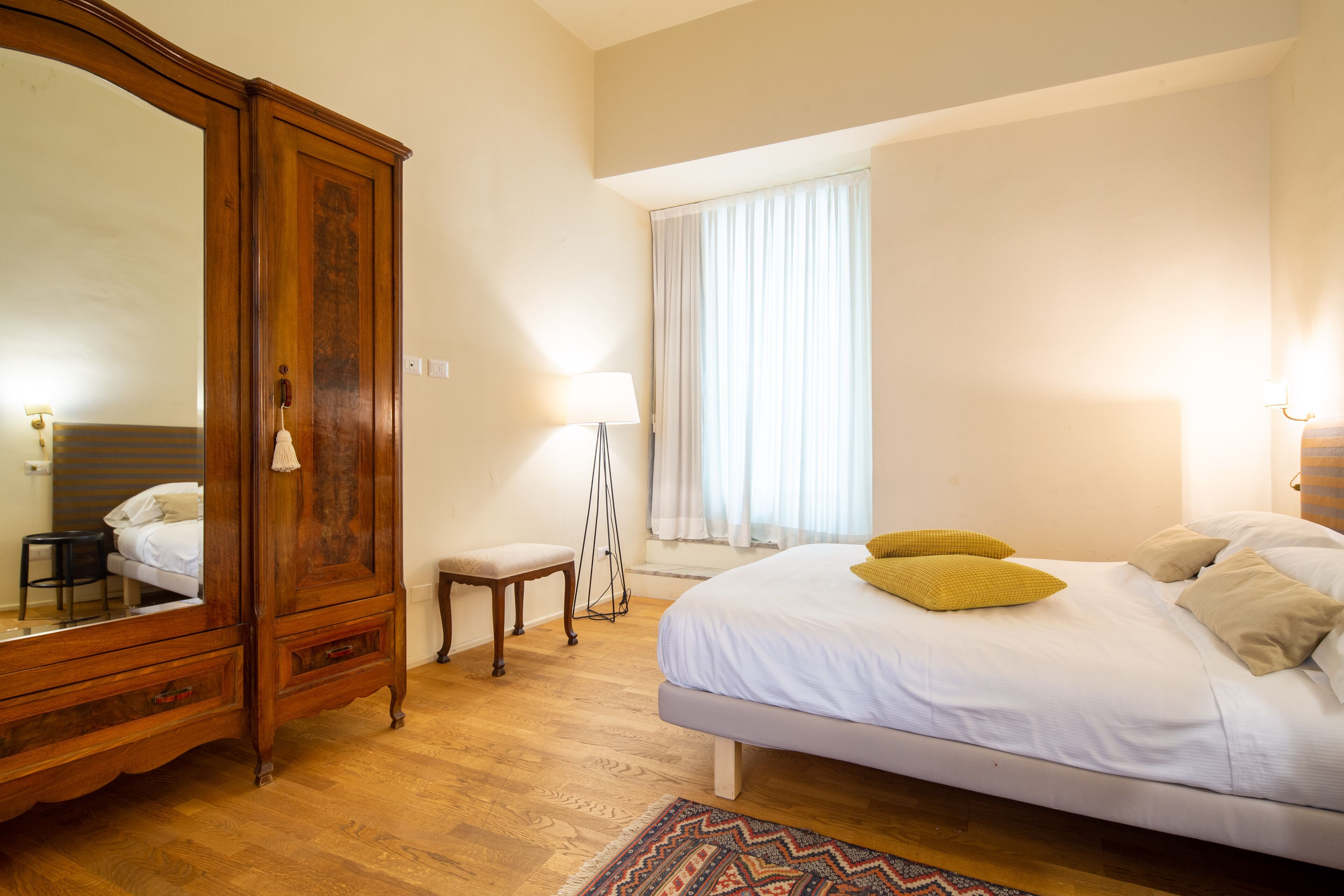 A bedroom with a wooden wardrobe, a bedside table, a lamp, a bed with pillows, and a window with curtains.