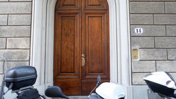 Puerta doble de madera con manija de latón, flanqueada por dos patinetes, y una pared de ladrillo con placa de número.
