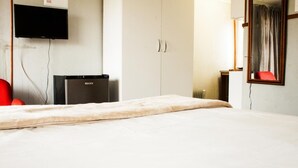 A hotel room with a bed, a television mounted on the wall, a wardrobe, and a small desk with a chair.