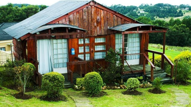 A rustic wooden cabin with a porch, surrounded by well-maintained gardens and a scenic view of rolling hills.