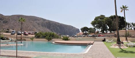 A swimming pool with a view of a mountain, surrounded by palm trees and a parking lot.