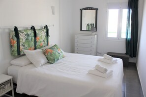 Un lit bien fait, avec du linge de lit blanc et deux oreillers, une commode blanche avec un miroir et une fenêtre avec des rideaux.