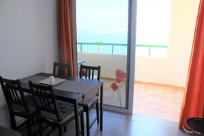 Un coin repas avec une table et des chaises, une vue sur la mer par une porte vitrée et un vase avec une fleur rouge posé sur le rebord de la fenêtre.