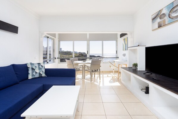 A modern living room with a blue sofa, a white coffee table, and a dining area with a table and chairs. There is a large TV on a wooden stand and a painting on the wall.