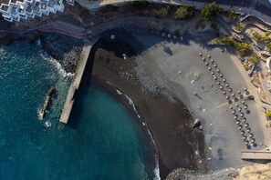 An aerial view of a coastal area with a rocky beach, a pier, and a promenade with palm trees.