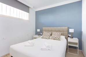 A neatly made bed with two pillows, a bedside table with a lamp, and a window with blinds.