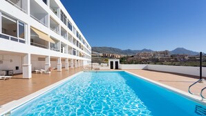 A swimming pool with a clear blue water surface, surrounded by a white building with multiple balconies and a view of mountains in the background. - Tranquilidad, playa y vistas al mar (Callao Salvaje)