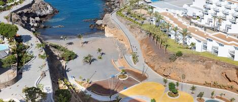 An aerial view of a coastal resort with a beach, palm trees, and a white building complex.