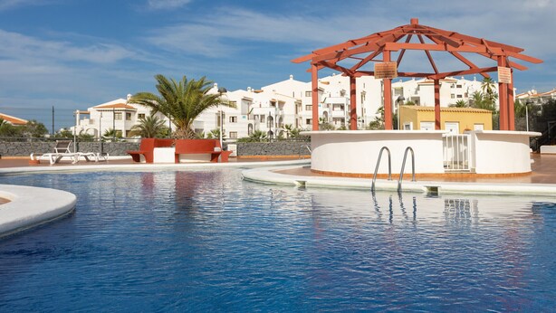 Una piscina con un pabellón, palmeras y edificios blancos al fondo.