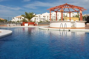 A swimming pool with a gazebo, palm trees, and white buildings in the background.