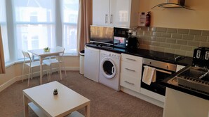 Ett kompakt kök med tvättmaskin, mikrovågsugn och brödrost. Det finns ett litet bord med en stol och ett fönster med gardiner.