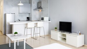 Cocina moderna con microondas integrado, refrigerador y televisor sobre un mueble blanco. Hay dos taburetes de bar y una pequeña mesa con una planta.