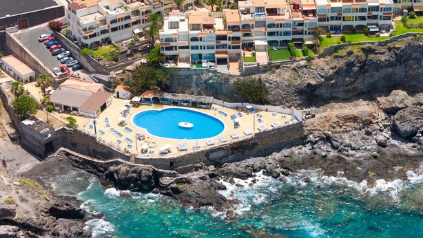 Un hotel frente al mar con una piscina circular rodeada de sillas de descanso, situado en una costa rocosa.