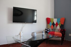 Uma sala de estar moderna com uma TV de tela plana na parede, uma mesa de centro de vidro, uma poltrona colorida e um pequeno vaso branco.