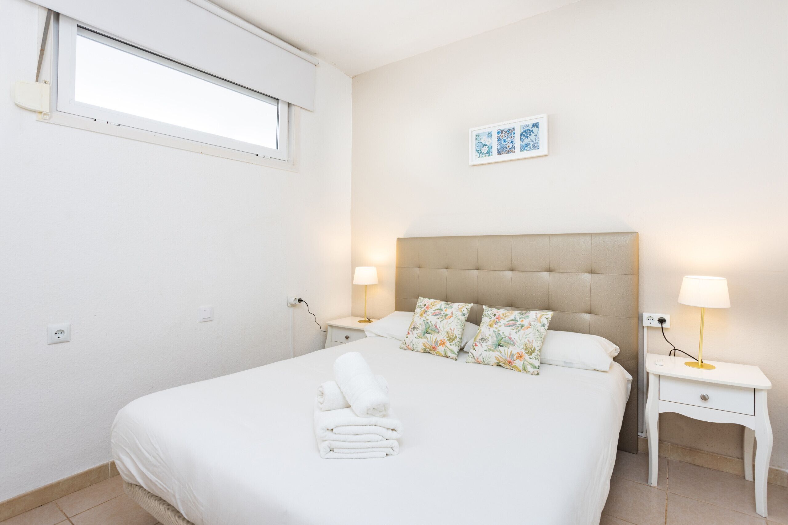 A bedroom with a bed, two bedside tables, a window, and a wall-mounted picture.
