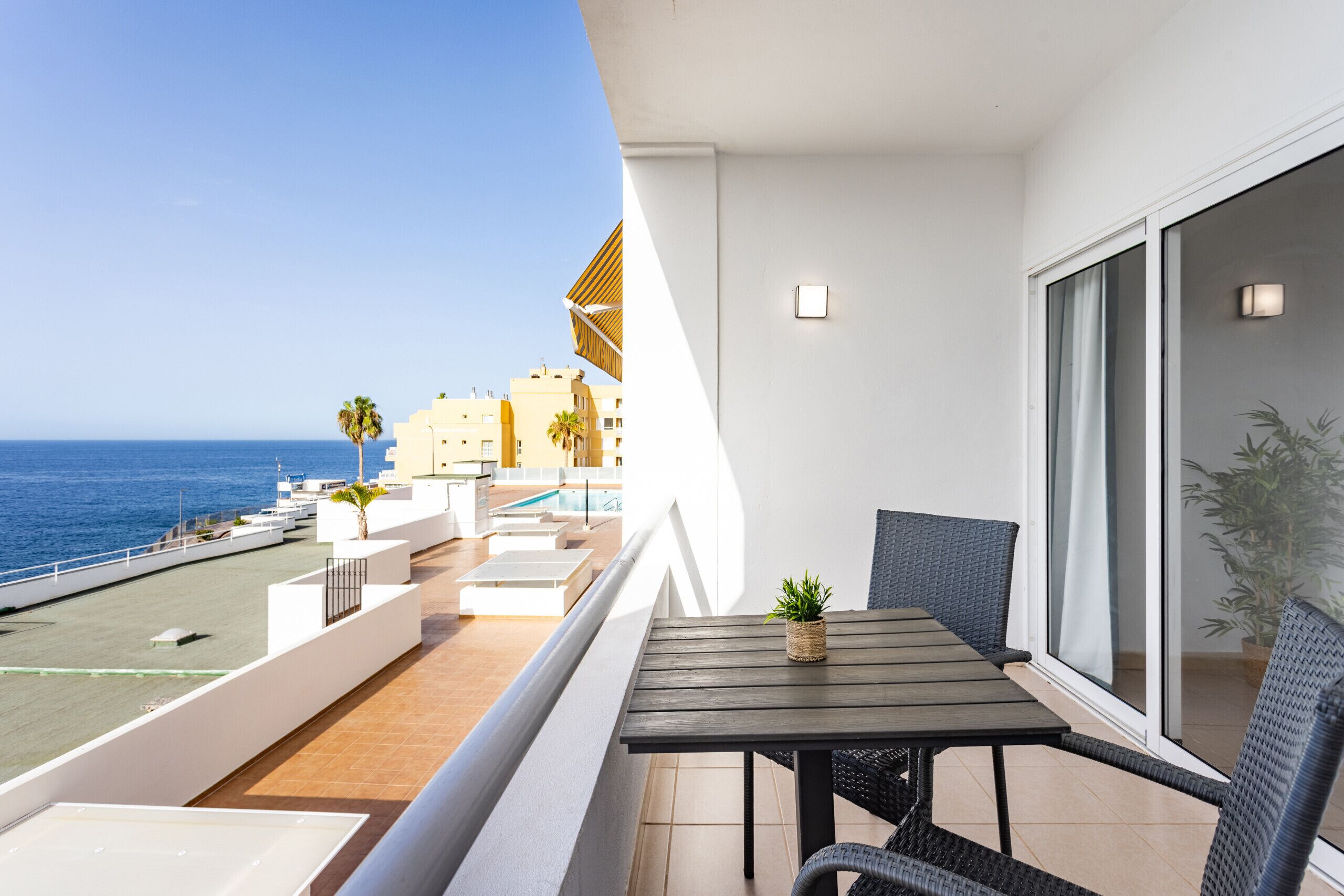 A balcony with a table and chairs, overlooking a pool and the sea, with buildings in the background.