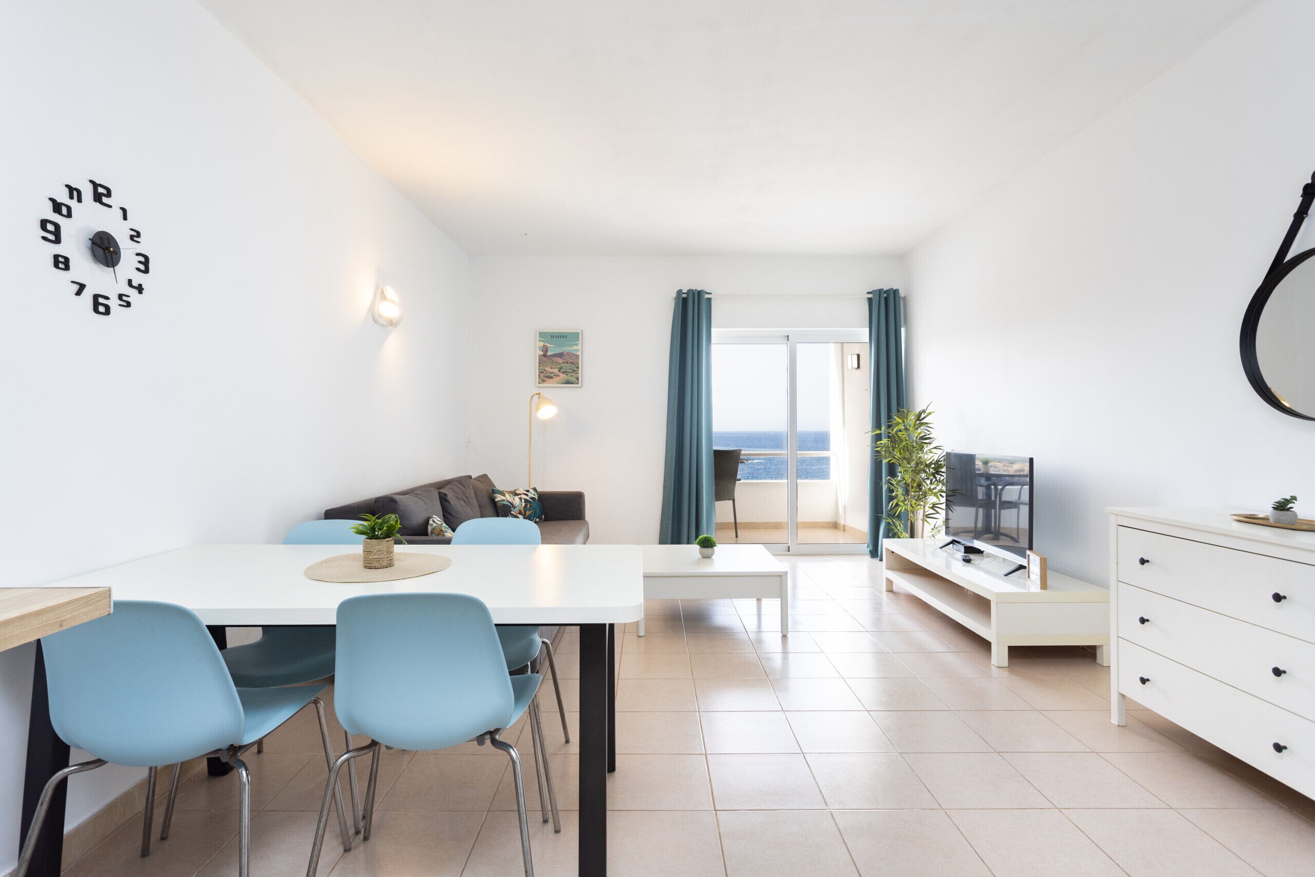 A modern living room with a dining area, a sofa, a TV, and a view of the sea.