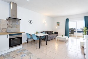 Una cocina moderna con horno y estufa integrados, un comedor con mesa blanca y sillas azules, un sofá gris y vistas al mar a través de amplias ventanas.