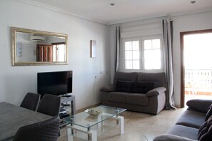 A living room with a flat-screen TV, a glass coffee table, a sofa, and a dining area with chairs.
