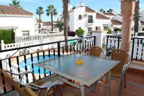 A terrace with a glass table and wicker chairs, overlooking a swimming pool and a white building with a tiled roof. - 4 Bed Fantastic Apt, Private Pool Close to Golf, Beach and Entertainment (Orihuela Costa)