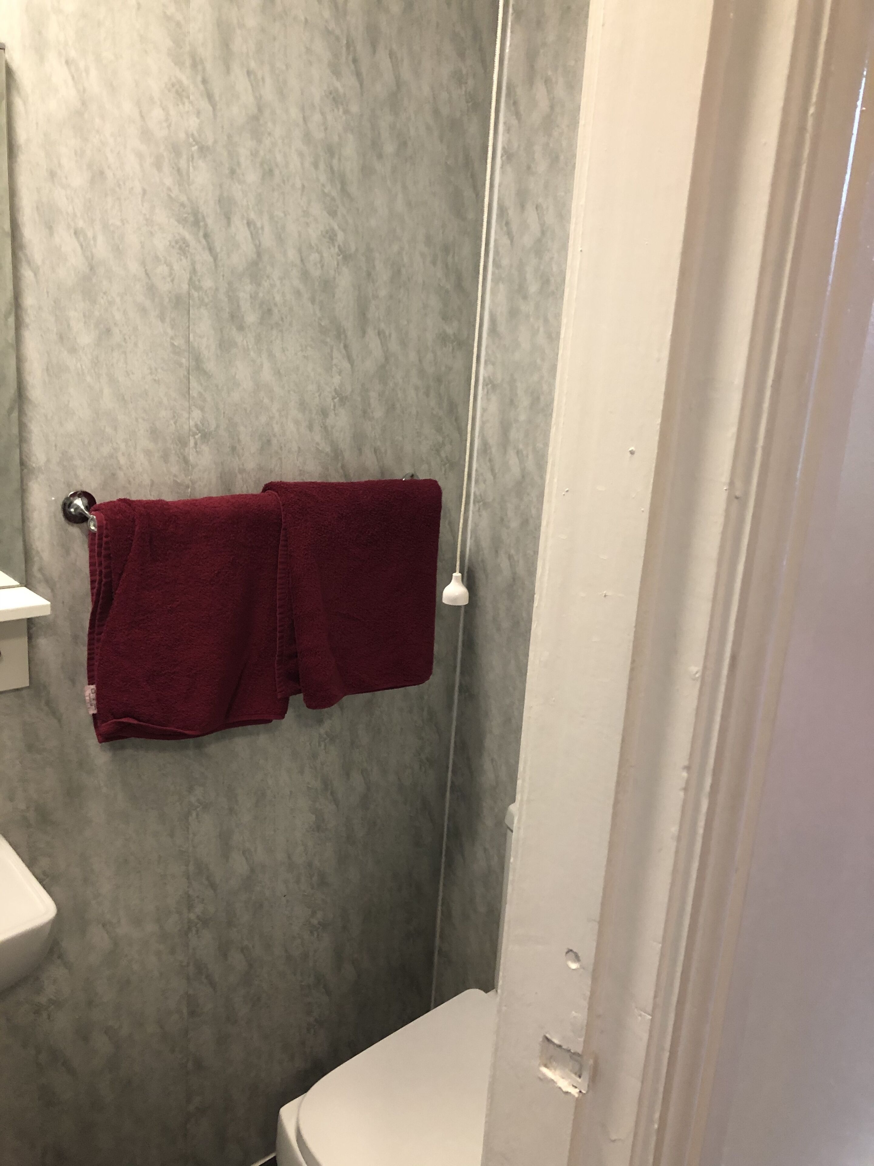 Kylpyhuoneessa on harmaa, kuvioitu seinä, valkoinen wc-istuin ja punainen pyyhe, joka on ripustettu metalliseen pyyhetelineeseen.