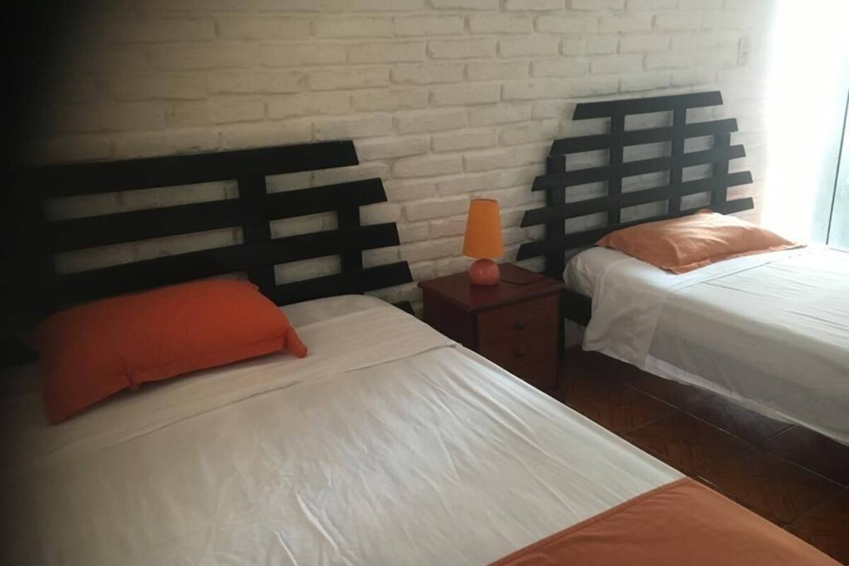 Dos camas individuales con ropa de cama blanca y naranja, un cabecero de madera, una mesita de noche con lámpara y una pared de ladrillo blanco.