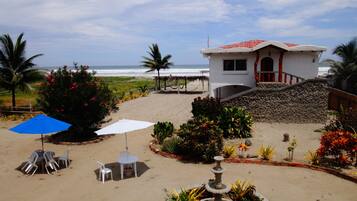 Una propiedad frente a la playa con un edificio blanco, una escalera de piedra, un área de comedor con sillas y mesas, y una pequeña fuente.