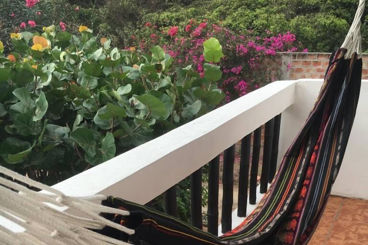 Una hamaca en un balcón con barandilla blanca, rodeada de follaje verde exuberante y flores rosadas.