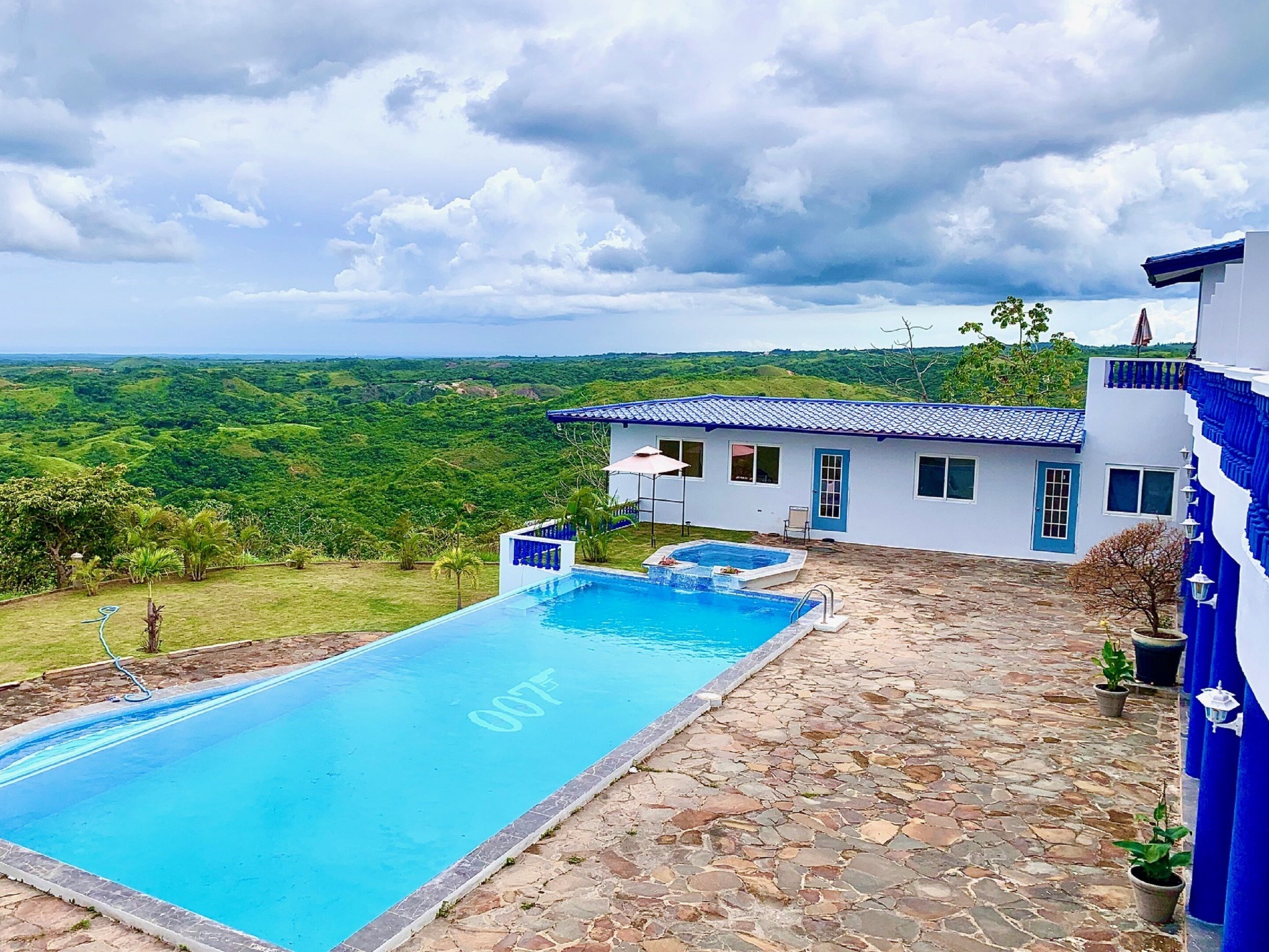 A pool area with a stone patio, a house with a blue roof, and a lush green landscape in the background.