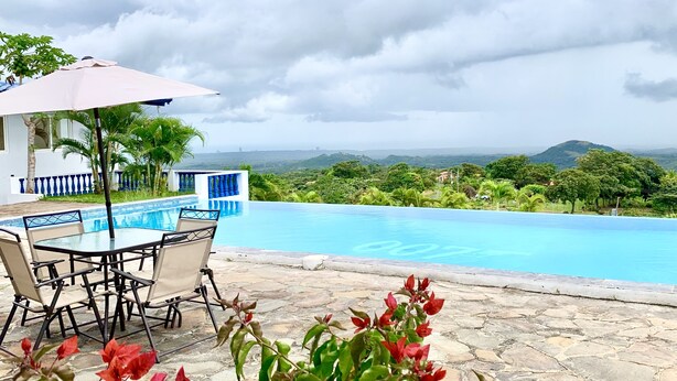 Zona de piscina con mesa y sillas, vista a un paisaje exuberante y cielo despejado.
