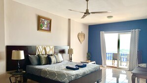Habitación de hotel con una cama grande, ventilador de techo, balcón con vistas y un adorno decorativo en la pared.