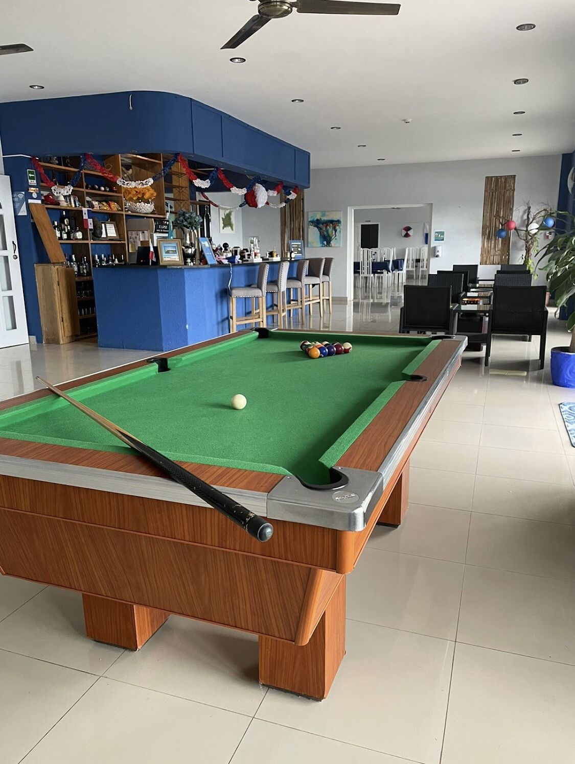 A pool table with a pool cue and balls, a bar area with shelves and chairs, and a seating area with tables and chairs.