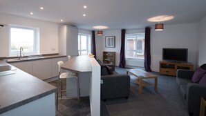 A modern apartment with a kitchen, dining area, and living room. It includes a bar area, a sofa, a TV, and a bookshelf.
