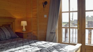 A wooden cabin room with a bed, a nightstand with a lamp, and a window with a view of the outside. - Baytree Lodge - UK13322 (Melbourne, near Derby)