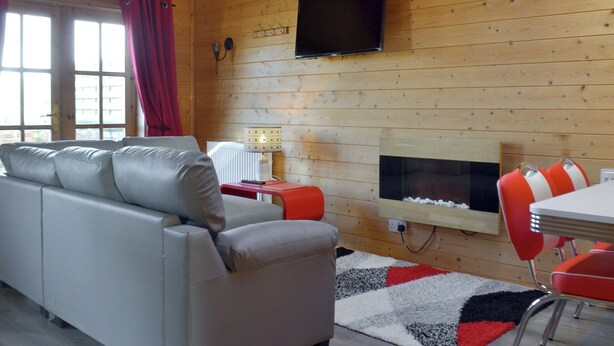 A wooden cabin interior with a sofa, a TV mounted on the wall, a fireplace, and a dining area with red chairs. - Baytree Lodge - UK13322 (Melbourne, near Derby)