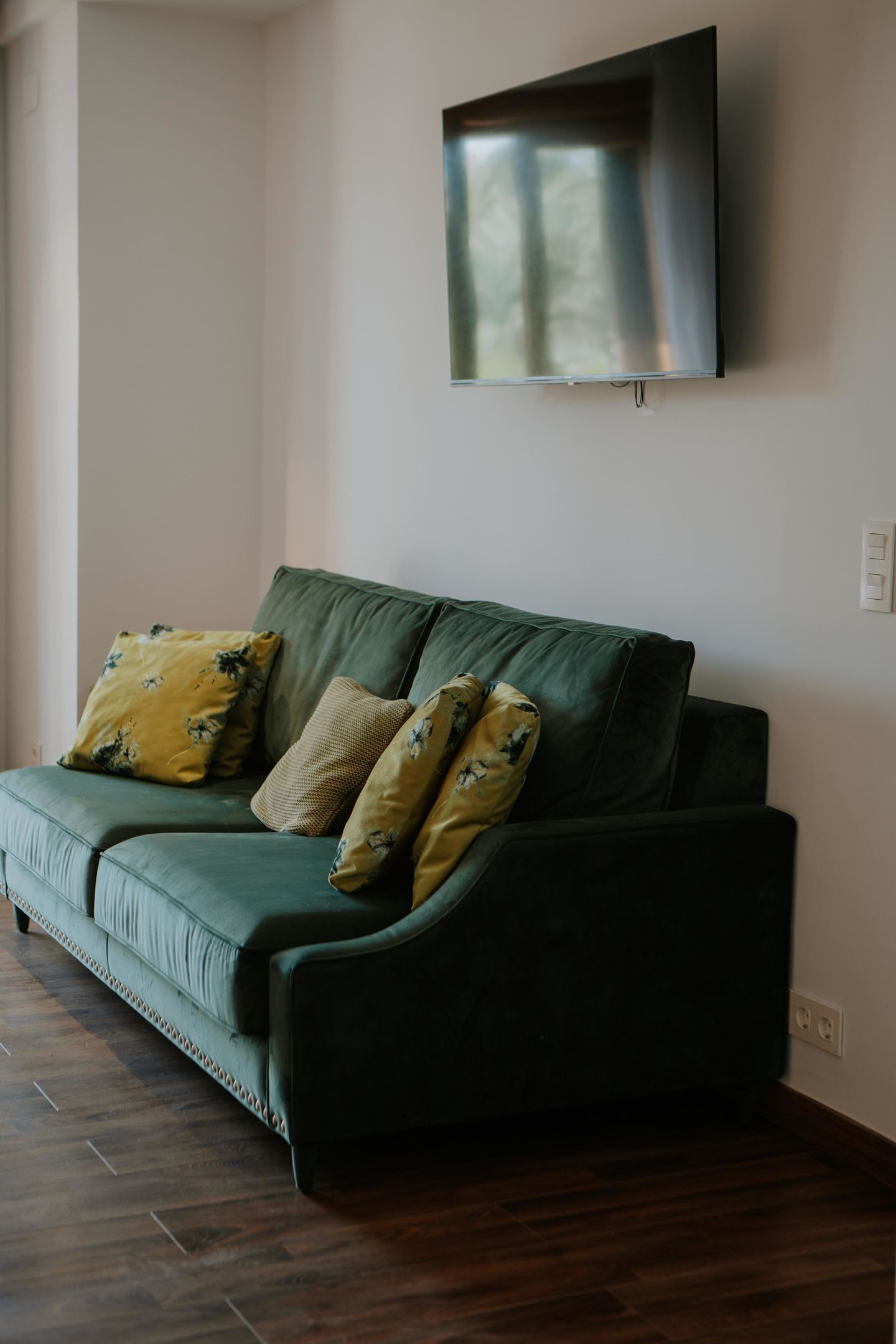 A green sofa with floral cushions, a flat-screen TV mounted on the wall, and a wooden floor.