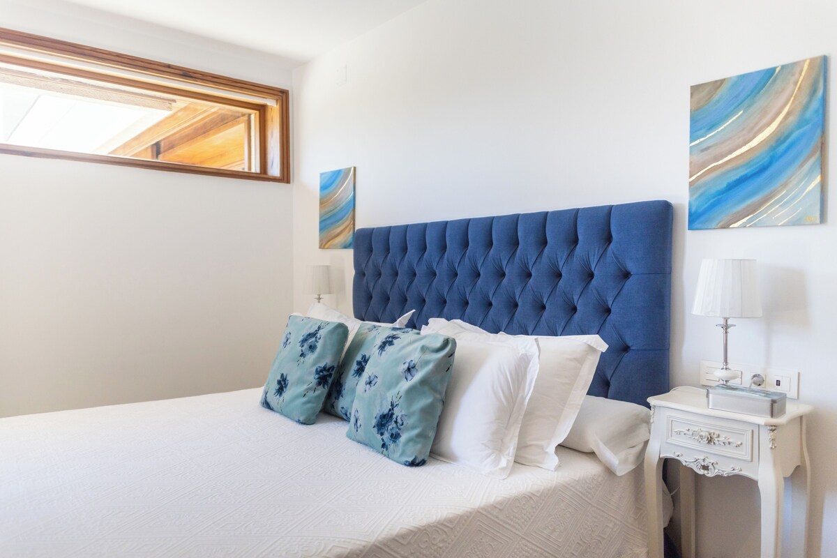 A bedroom with a large bed, a tufted headboard, floral pillows, a bedside table with a lamp, and abstract wall art.