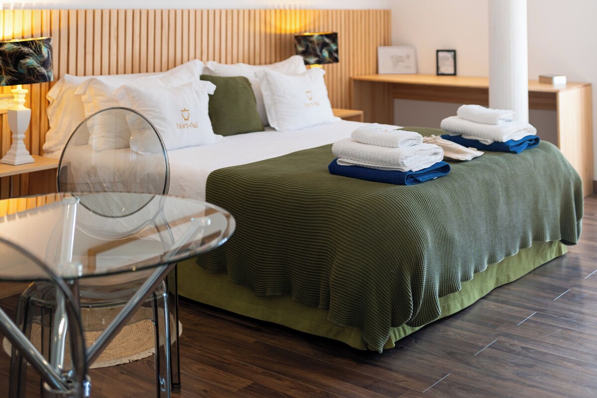 A hotel room with a bed, a glass table, a chair, a green bedspread, and a framed picture on the wall.