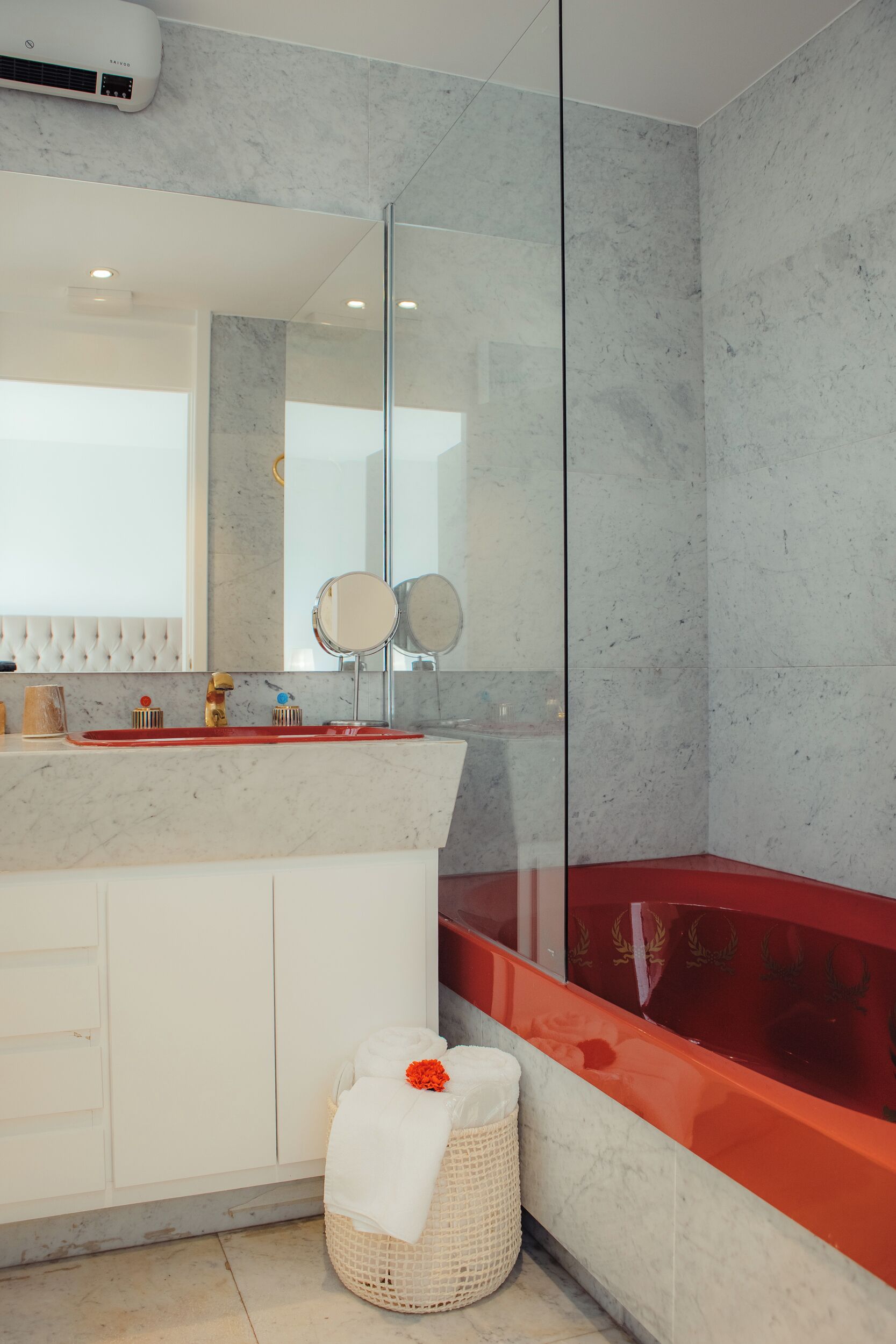 Un baño moderno con lavamanos de mármol, dos espejos, una bañera roja y una cesta blanca para toallas.