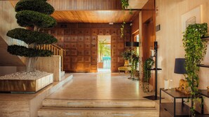 Un interior de madera con una escalera que lleva a una puerta, un árbol en maceta y una lámpara sobre una mesa.