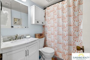 Une salle de bain avec un meuble-lavabo blanc, un évier et des toilettes. Il y a un rideau de douche orné d’un motif en forme de soleil rayonnant.