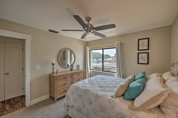 Une chambre à coucher comprenant un lit, un ventilateur de plafond, une commode, une fenêtre avec des rideaux et des œuvres d’art encadrées accrochées au mur.