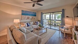 A spacious living room with a ceiling fan, a large window with a view of the outdoors, a sofa set, and a coffee table.